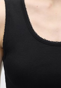 Gros plan d'une personne portant un débardeur noir à côtes avec une bordure en dentelle le long de l'encolure et de l'emmanchure.