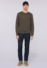 Olivgrön sweatshirt med rund halsringning, tillsammans med mörk denimjeans och bruna sneakers. Har en subtil logotyp på bröstet.
