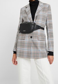 Borse a marsupio in pelle nera fissata in vita, abbinata a un blazer a quadri nei toni del blu, marrone e bianco, completata da pantaloni bianchi.