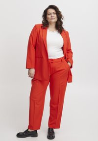 Kirkas punainen muotoiltu puku, jossa on löysä bleiseri ja yhteensopivat housut, yhdistetty valkoiseen paitaan. Mallilla on päällään mustat loafers-kengät. Sujuva kangassekoitus.