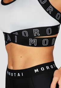 Sujetador deportivo en blanco y negro que presenta una banda negra ancha con "MOROTAI" en letras blancas. Tejido suave con costuras en contraste y diseño de espalda nadadora.