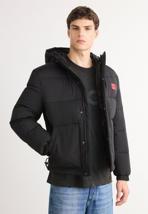 Black puffer jacket with a hood, zip closure, stitched panels, and a small red logo patch. Features elastic cuffs and adjustable hem.
