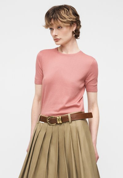 Γυναίκα με κοντά κυματιστά μαλλιά, φοράει χρυσούς κρίκους σκουλαρίκια, ροζ μπλούζα με κοντά μανίκια, πλισέ μπεζ φούστα και καφέ ζώνη με χρυσή αγκράφα.