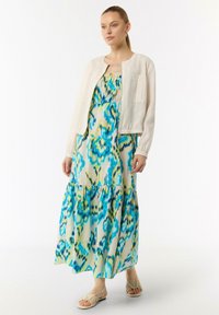 Maxi sukienka w niebiesko-zielonym abstrakcyjnym wzorze z warstwowym krojem, połączona z kremową krótką kurtką i płaskimi sandałami.