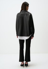 Camicia oversize in finta pelle nera con spalle cadenti, colletto alto e orlo bianco a strati, abbinata a pantaloni neri svasati e tacchi.