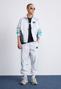 Bílý a šedý teplákový komplet s tyrkysovými akcenty, obsahující zipovou bundu a odpovídající kalhoty, doplněný o šedé tenisky a sluneční brýle.