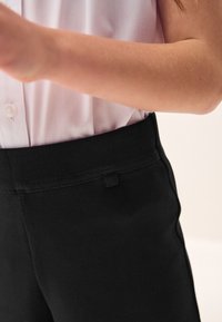 Musta joustavat housut, joissa on sileä vyötärönauha ja pieni lenkkidetalji. Kangas on pehmeää ja siinä on matta pinta. Vaaleanpunainen paita näkyy osittain.