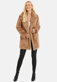 Manteau en fausse fourrure beige avec un grand col, coupe surdimensionnée, boutons à l'avant et poches latérales, associé à des legging en cuir noirs et des bottines à talons.