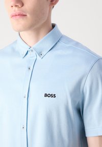 Šviesiai mėlyna polo marškinėlių su tekstūruotu audiniu ir mygtukų apykakle. Kairėje krūtinės pusėje yra juodas "BOSS" logotipas.
