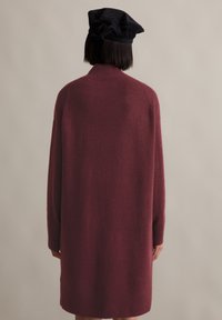 Person, der mit dem Rücken zur Kamera steht, trägt ein langes, burgunderfarbenes Sweater-Kleid und eine schwarze, weiche Mütze vor einem schlichten beige Hintergrund.