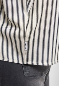 Tela a rayas en tonos crema y negro, con una etiqueta en el dobladillo. Jean gris visible con una sutil textura y detalles de costura.