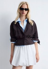 Black cropped jacket made of linen over a light blue button-up shirt, paired with a white, textured, voluminous skirt. Black sunglasses included.
