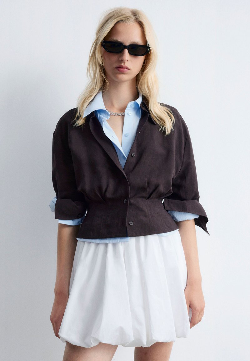 Black cropped jacket made of linen over a light blue button-up shirt, paired with a white, textured, voluminous skirt. Black sunglasses included.