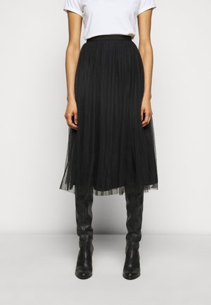 Vrouw die een zwarte geplooide midirok, een wit T-shirt met korte mouwen en zwarte leren laarzen tot over de knie draagt, staand tegen een effen achtergrond.