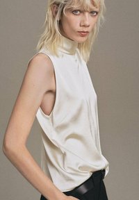 White sleeveless top with a high neckline, made of smooth fabric, featuring side slits and a relaxed fit, styled with black pants.