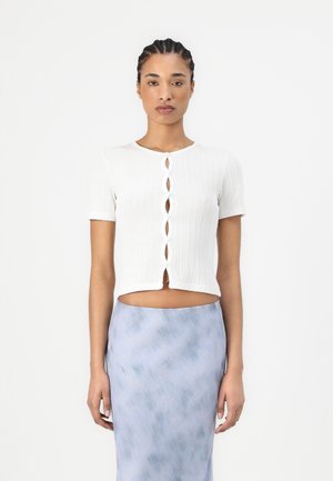 White ribbed short-sleeve crop top with a button closure featuring an elongated loop design. Paired with a light blue, smooth, patterned skirt.