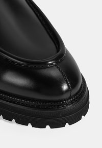 Botte cheville en cuir noir avec une pointe arrondie, finition brillante et semelle en caoutchouc texturée. Présente des détails cousus le long du bord.