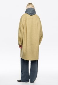 Persona vista desde atrás que lleva un abrigo de lana beige sobre una sudadera con capucha gris oscuro y pantalones oscuros holgados con zapatos de punta.