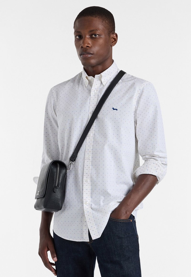 Biała koszula z guzikami, pokryta kolorowymi kropkami, z małym niebieskim logo, zestawiona z ciemnymi dżinsami i czarną torbą na ramię.