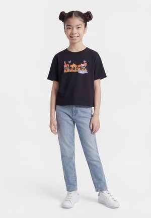 Pige med dobbelte knolde iført en sort T-shirt med blomsterprint, lyseblå jeans og hvide sneakers, stående og smilende på en hvid baggrund.