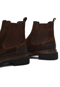 Bruine suede enkellaarzen met ribbel-elastische zijkanten, een contrasterende donkerbruine leren zool, en handvatten aan de achterkant voor eenvoudig aan- en uittrekken.