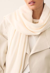 Cream scarf made of soft fabric, draping loosely around the neck; paired with a light beige trench coat featuring detailed stitching.