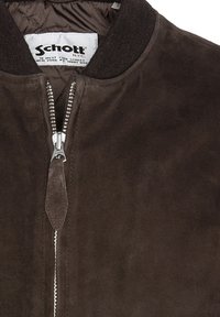 Giacca bomber in suede marrone con collo a coste, zip frontale e fodera trapuntata. Presenta un'etichetta con logo all'interno. Superficie testurizzata.
