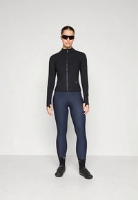 Egy nő fekete hosszúujjú kerékpáros pólót, tengerészkék párnázott kerékpáros leggingset, fekete zoknit, kerékpáros cipőt és sötét napszemüveget visel, egy fehér háttér előtt állva.