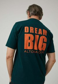 Camiseta de algodón de color verde oscuro con un gran texto naranja en la espalda que dice "DREAM BIG ALTONALOCK". Cuello redondo y mangas cortas.