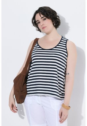 Frau mit kurzen lockigen Haaren, die ein schwarz-weiß gestreiftes Tanktop, weiße Hose, braune Umhängetasche und ein hellbraunes Armband trägt, steht vor einer weißen Wand.