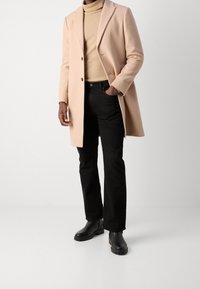 Abrigo de mezcla de lana beige con un cierre de botón único, combinado con un suéter de cuello alto beige claro y jeans negros de pierna recta. Botas de cuero negras.