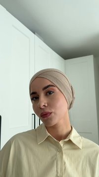 Donna con un turbante beige e una camicia color giallo chiaro con colletto, con un piercing al naso, che posano all'interno contro armadietti bianchi.