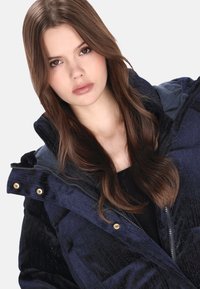 Granatowa kurtka puchowa o prążkowanej fakturze, z wyraźnym kołnierzem i złotymi elementami ozdobnymi. Pod kurtką widoczna czarna podkoszulka.