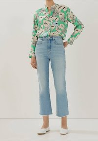 Blusa verde con motivo floreale e scollatura a V, abbinata a jeans azzurri alla caviglia e scarpe bianche senza lacci. Tessuto morbido, vestibilità rilassata.