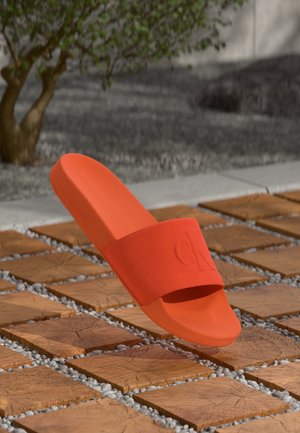 Une sandale slide orange unique avec un logo embossé sur la bride reposant sur un sol en dalles de bois avec de petits cailloux gris entre les dalles.