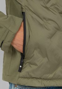 Oliwkowa kurtka puchowa z teksturowanego materiału, boczna kieszeń na zamek błyskawiczny oraz czarny kontrastowy zamek. Miękki, ocieplany materiał o gładkim wykończeniu.