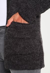 Ciemnoszara sweterka z dzianiny, charakteryzująca się teksturowanym, puszystym materiałem, z widoczną przednią kieszenią i luźnym krojem.