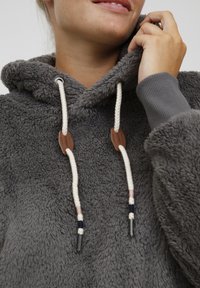 Hoodie in fleece grigio con superficie testurizzata, dotato di un cordoncino con estremità in metallo e dettagli in pelle, e polsini a coste.