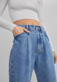 Jeans de mezclilla azul de tiro alto con pliegues en el frente, bolsillos laterales y un ligero desgaste. Top blanco de manga larga acortada con textura ribeteada.