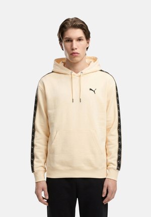 Joven con sudadera con capucha color crema con franjas negras con el logo en las mangas y pantalones negros, de pie frente a un fondo liso.