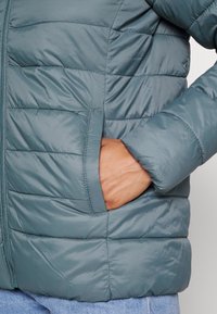 Casaco acolchoado em teal, com costuras horizontais, fecho frontal e bolsos laterais. O material é suave com um leve brilho.