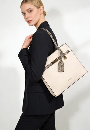Femme blonde en blazer noir portant un grand sac fourre-tout blanc avec des anses en cuir de serpent et des ferrures dorées sur l'épaule.