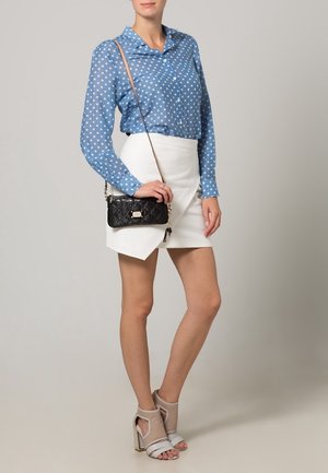 Kvinna som bär en blå prickig blus, vit asymmetrisk kjol, beige högklackade sandaler och håller en svart crossbody-väska.