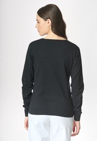 Una donna con lunghi capelli castani indossa un maglione nero, vista di spalle contro uno sfondo chiaro, che mette in risalto il design del capo.