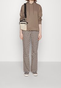 Brązowa bluza z kapturem, czarno-kremowe spodnie w kratę rozszerzane ku dołowi, czarno-białe buty w wzory, torba na ramię z kieszeniami na zamek. Stojąca w płaskiej pozycji.