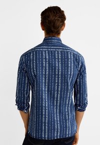 Camicia a buccia blu con motivo a righe verticali e forme astratte. Maniche lunghe arrotolate, colletto button-down e tessuto leggero.