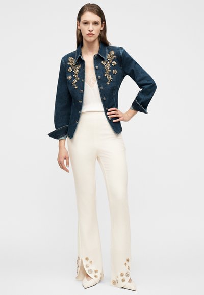 Γυναίκα φοράει σκουρομπλε τζιν μπουφάν με χρυσή κεντητή ανθική διακόσμηση, κρεμ τοπ και κρεμ παντελόνι ψηλόμεσο με αντίστοιχη κεντητή διακόσμηση στο τελείωμα.