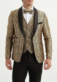 Blazer en velours doré à motifs paisley avec revers en satin noir, gilet assorti et nœud papillon, porté sur une chemise blanche, accompagné de pantalons noirs.