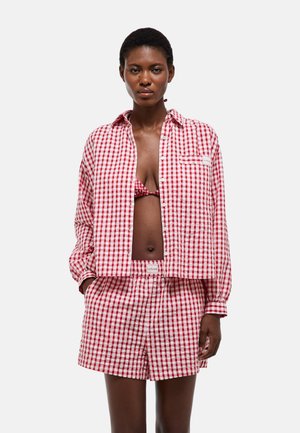 Femme portant une chemise à carreaux rouge et blanc, un short assorti et un haut de bikini rouge visible, debout devant un fond uni.