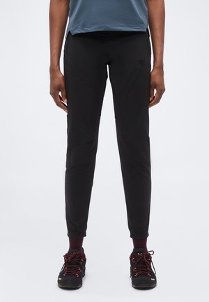 Osoba nosiaca čierne slim fit nohavice, bordové ponožky a čierne turistické topánky s červenými šnúrkami, stojaca na neutrálnom pozadí.